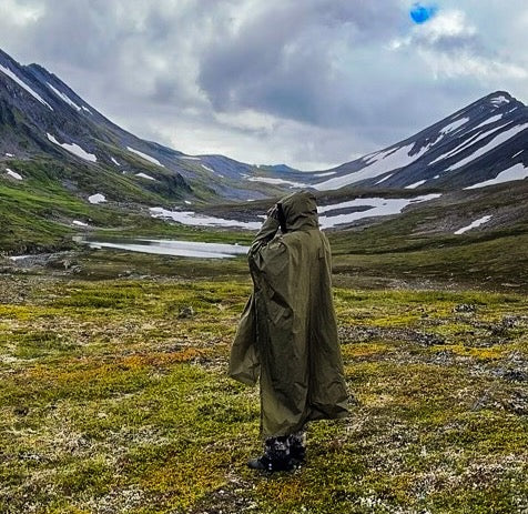 Person wearing a green Western Fly poncho standing in a mountainous landscape with a lake and snow patches.