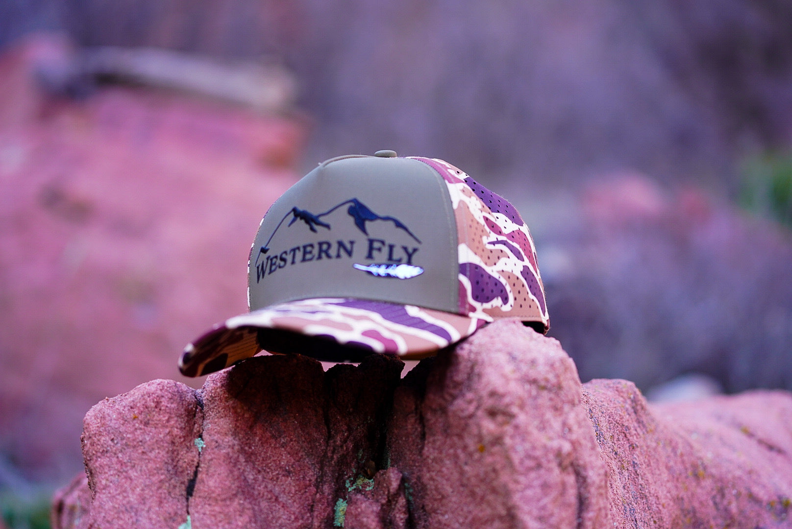 Camo-patterned cap with 'Western Fly' logo on a rock with a blurred natural background