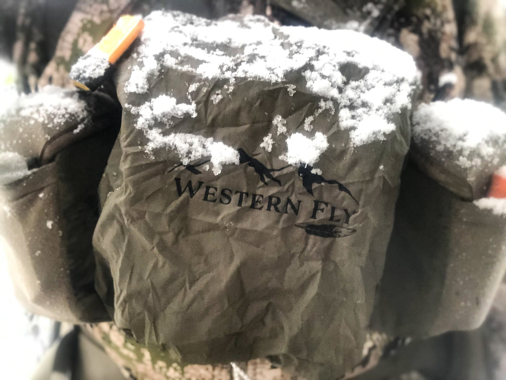 Snow-covered olive drab bino fly with 'Western Fly' logo on a blurred natural background
