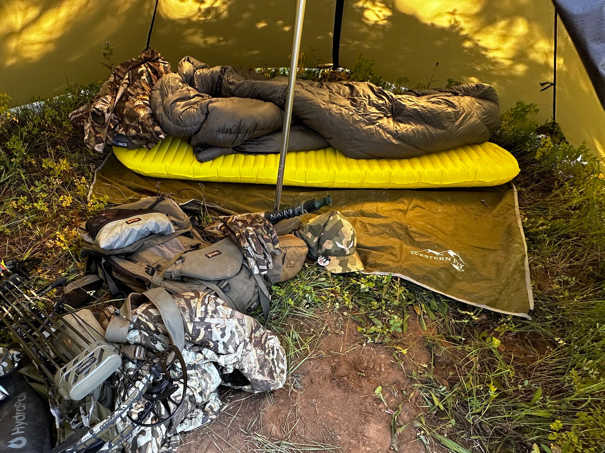 Sleeping bag on a yellow sleeping pad with camouflage gear nearby in a natural setting.